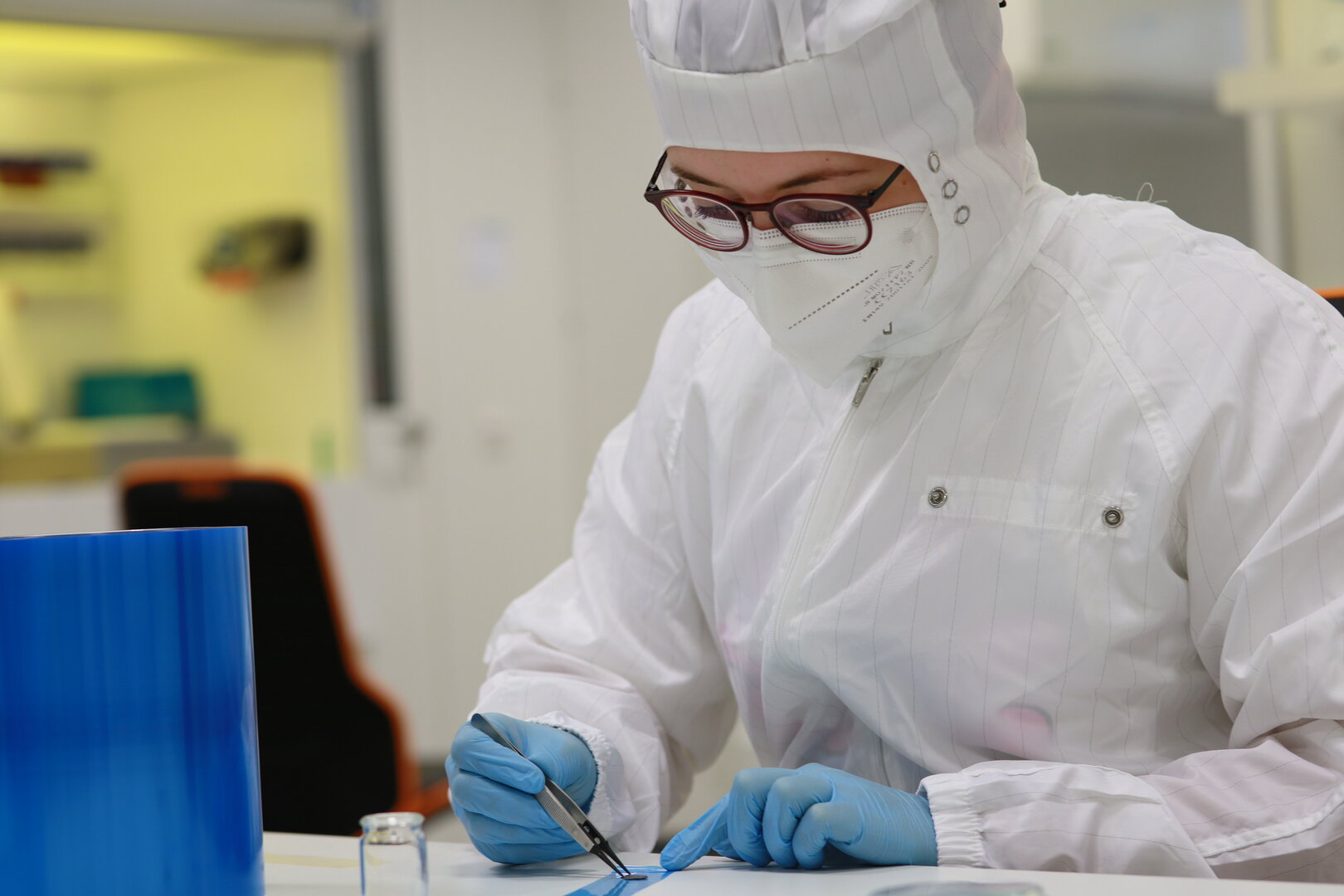 Eine Wissenschaftlerin in Schutzkleidung und mit FFP2-Maske zieht mit einer Pinzette ein Objekt von einem Klebefilm.